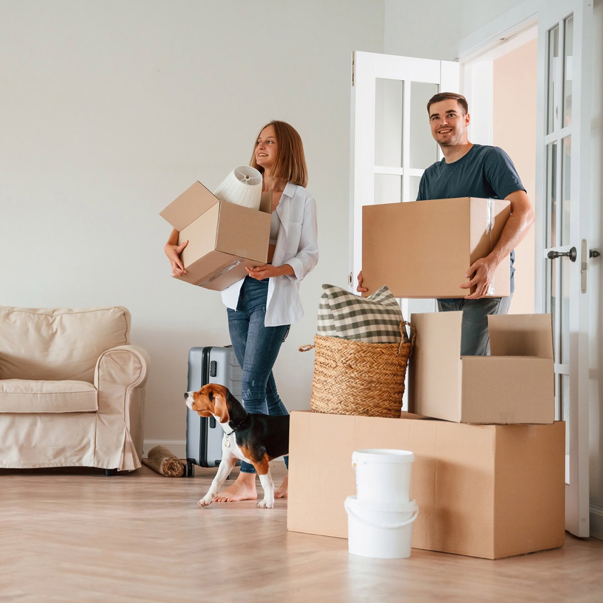 A woman and a man are walking into a new home carrying moving boxes with a dog.