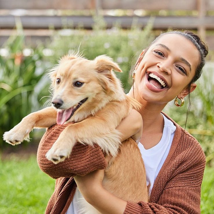 A young adult female sitting outside smiling while holding up a golden long-haired puppy